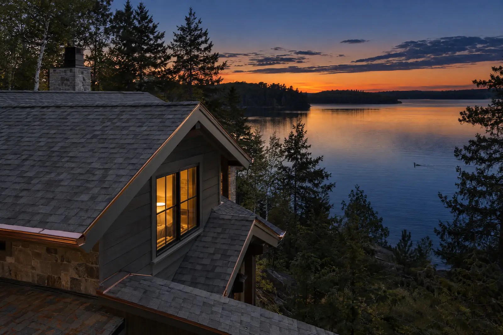 A shingled lake home above the water at dusk, with a loon on the lake