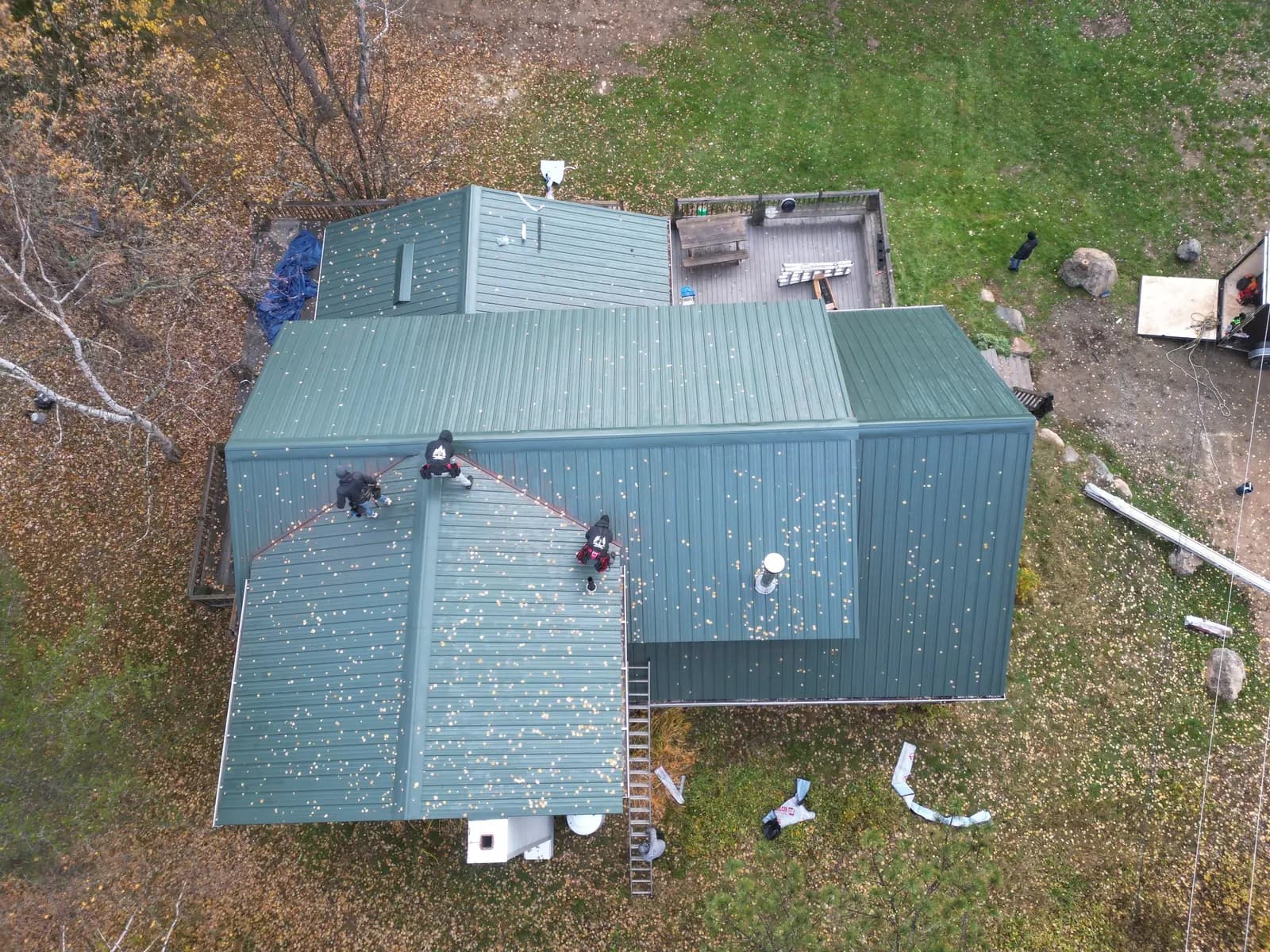 Aerial view of three crew members installing a sage-green standing seam roof on a large lodge, autumn leaves scattered on the panels