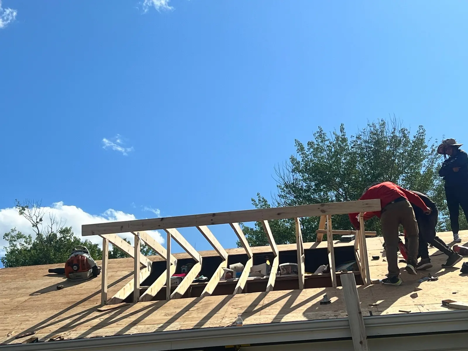 Crew framing a new roof dormer on a two-story home, dark siding, blue sky