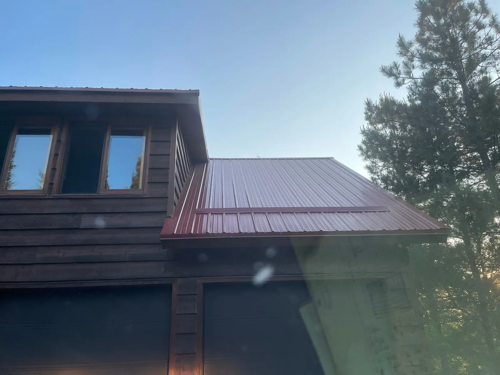 Dormer window on a dark log cabin with barn-red standing seam roof, car window reflection in frame