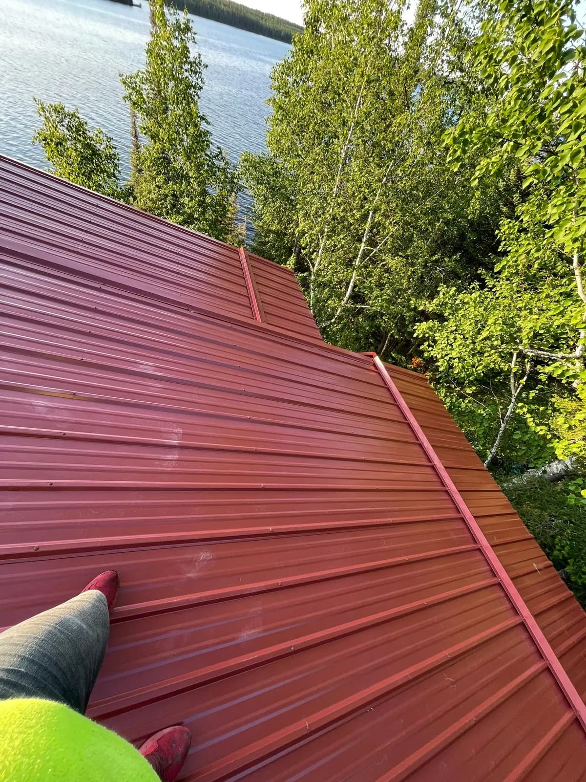 Point-of-view shot from a dark log cabin roof, barn-red standing seam panels, worker boots visible