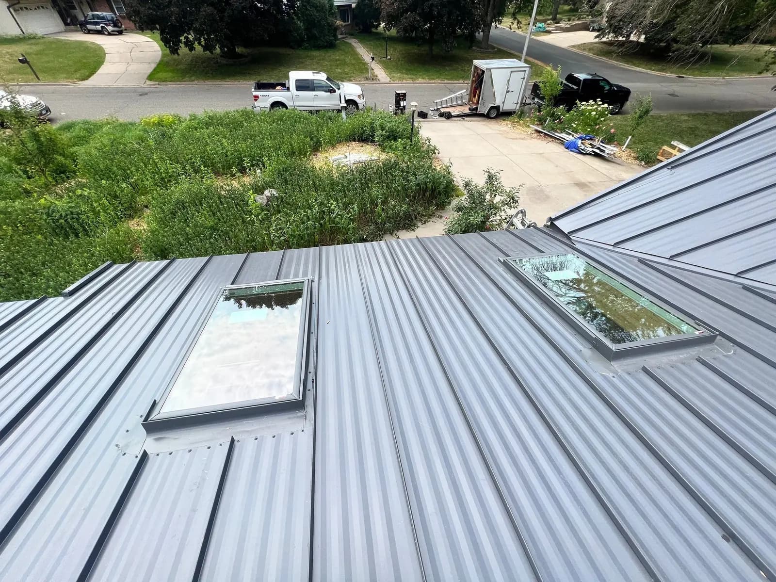 Looking down from a charcoal standing seam roof with skylights at a suburban street, work trailer and trucks parked below