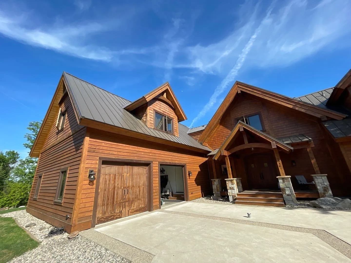 Large custom timber-frame home near Lake Vermilion, Minnesota with dark bronze standing seam metal roof, multiple gables and dormers, stone porch columns, blue sky, May 2023