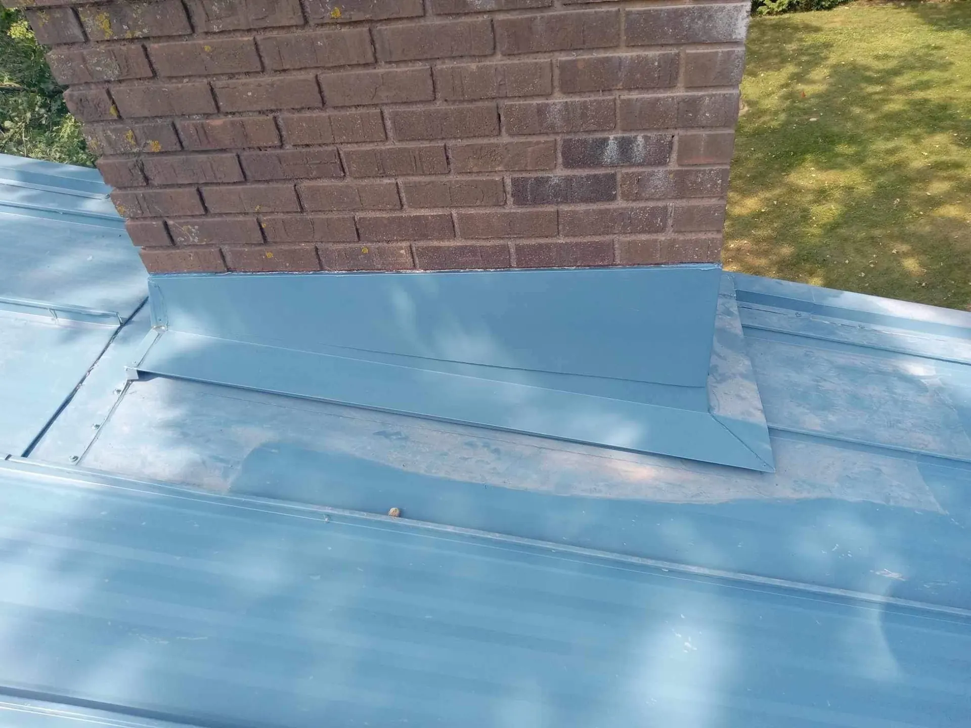 Powder-blue standing seam metal roof with brick chimney and new flashing on a Minnesota home, tree line in the background, August 2023