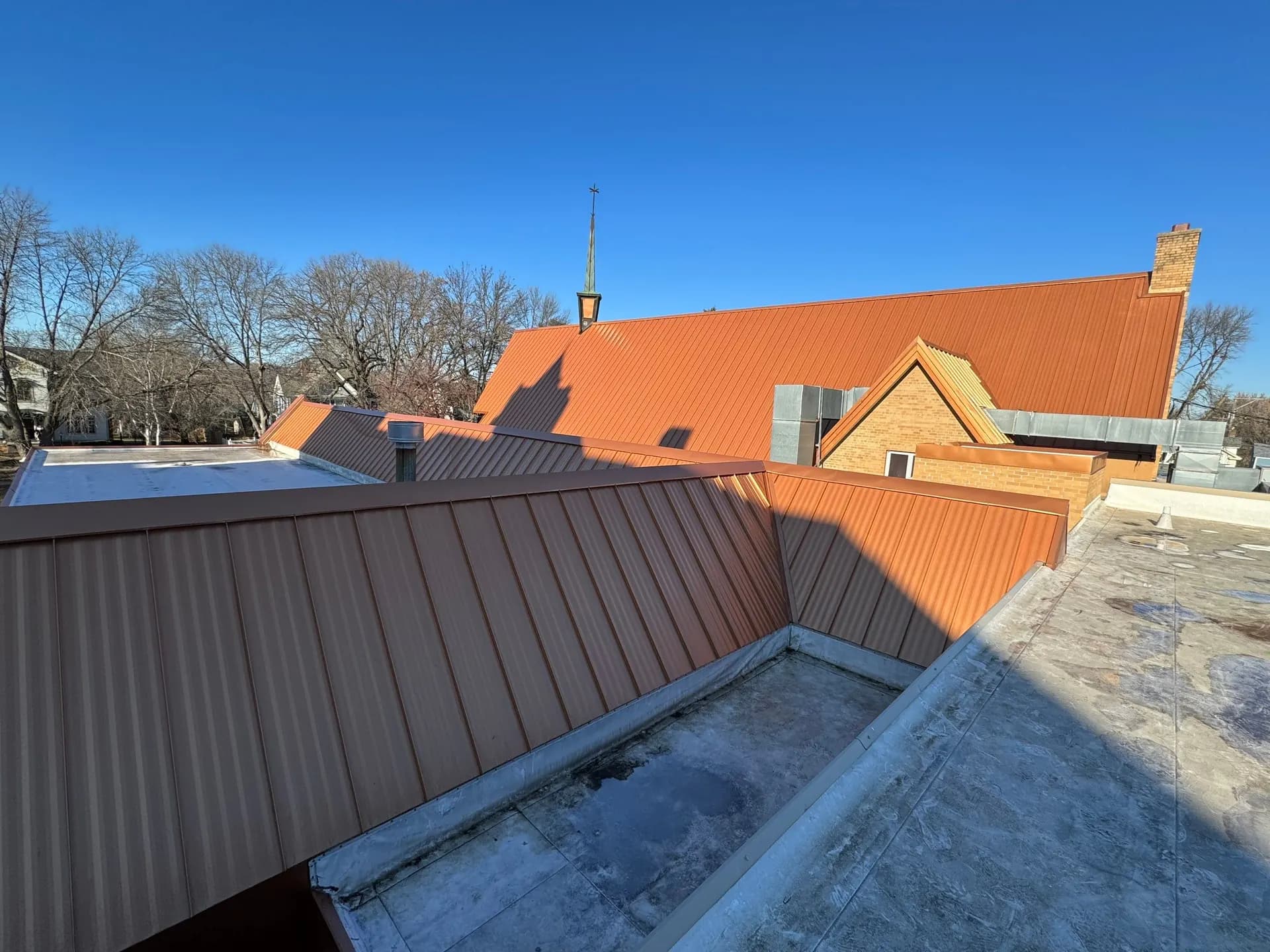 Copper-orange standing seam metal roof on a brick church with a steeple and cross, brick gable dormer, and galvanized rooftop ductwork, clear blue sky, November 2023