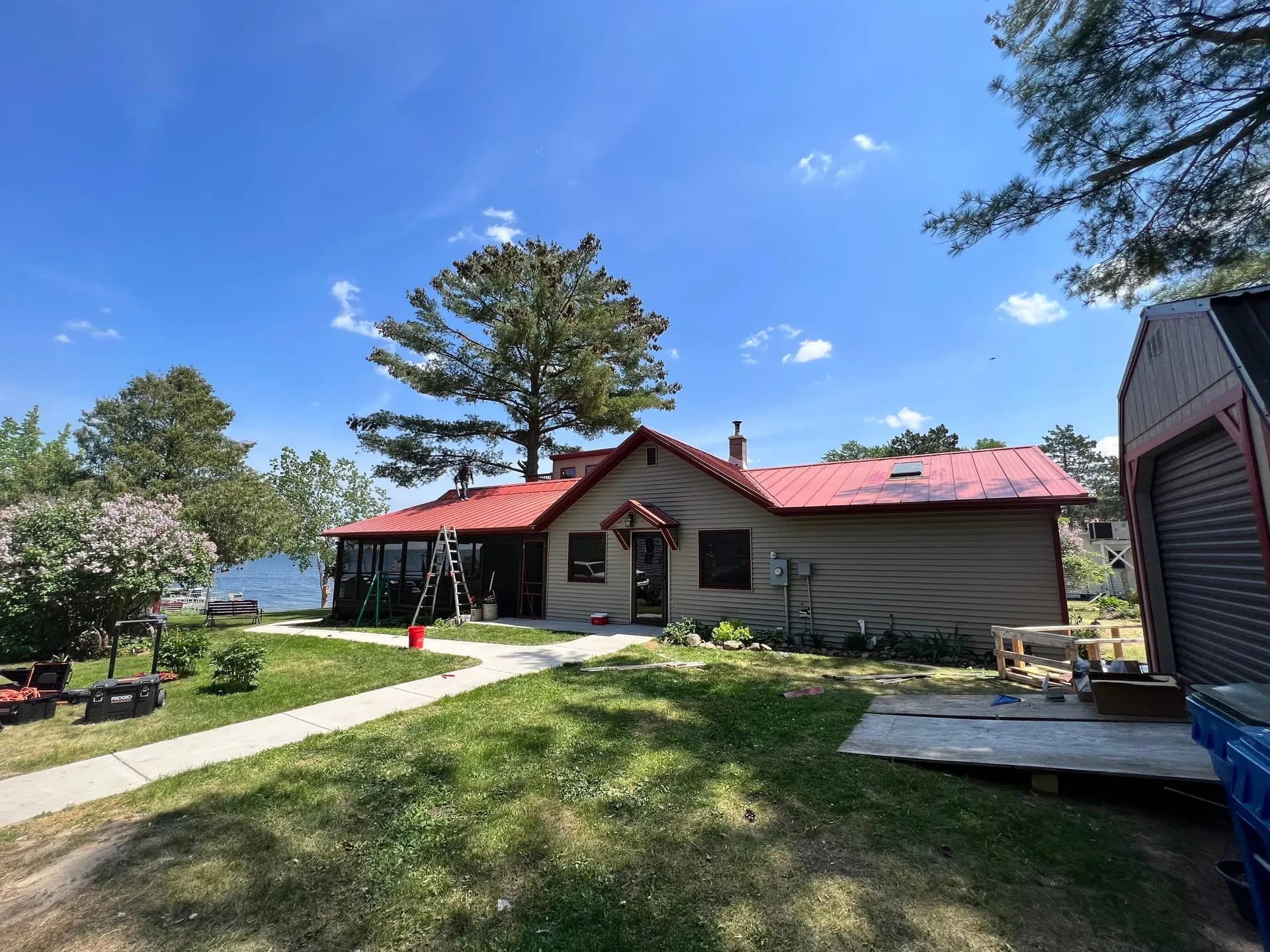 Lakefront ranch home with barn-red standing seam metal roof, screened porch, lilac bushes in bloom, lake and dock visible past the yard, crew member on the roof, May 2023