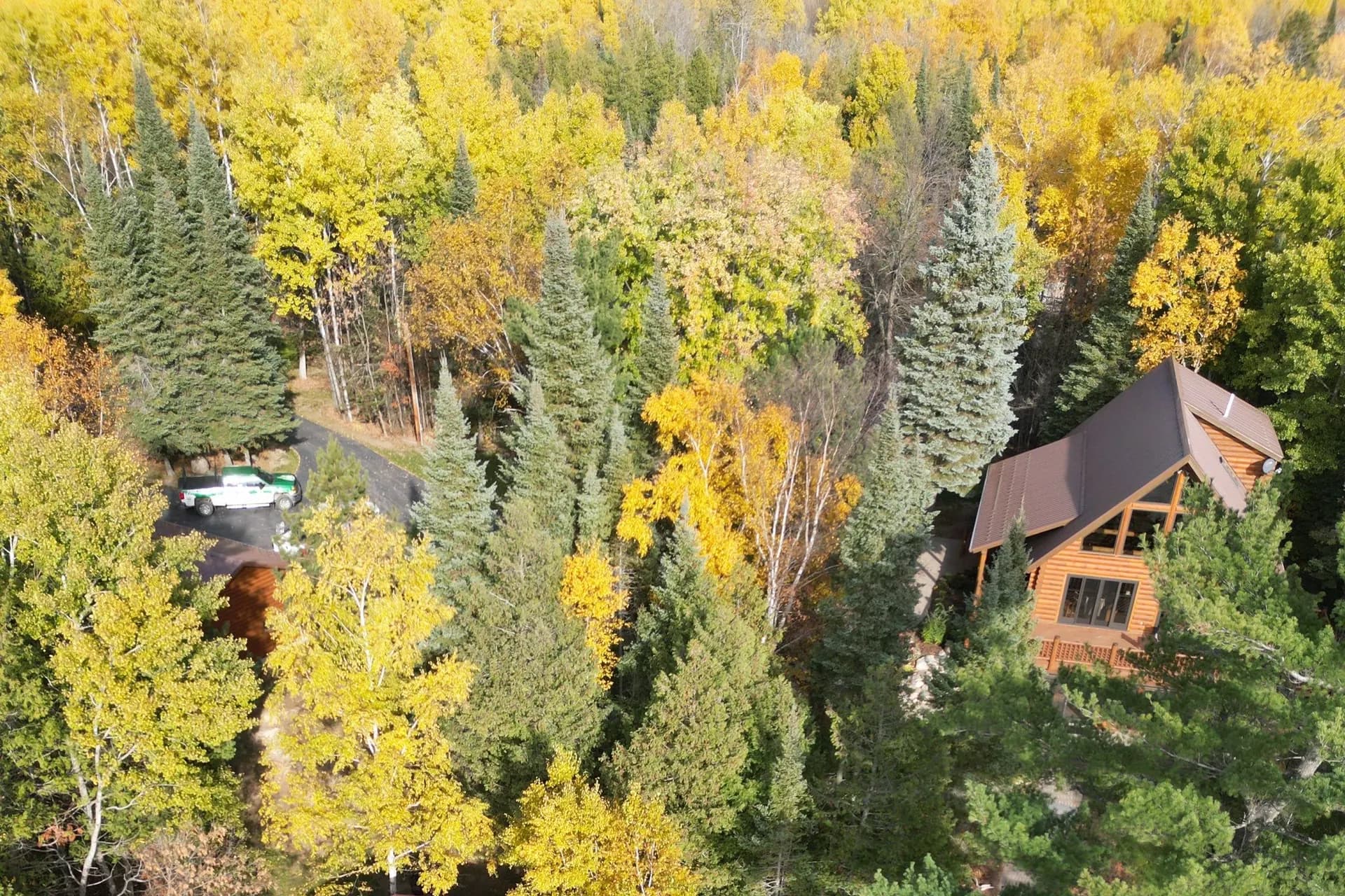 Aerial view of an A-frame timber lodge with brown standing seam metal roof in a northern Minnesota lake-country clearing, fall foliage, work truck parked in the driveway, October 2023
