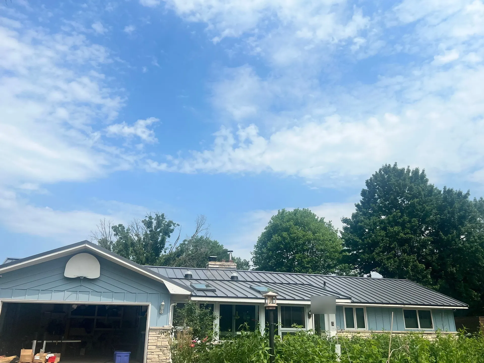 Light-blue suburban ranch home in the Twin Cities metro with new charcoal standing seam metal roof, attached garage, stone accents, trees and neighborhood street in the background, June 2023